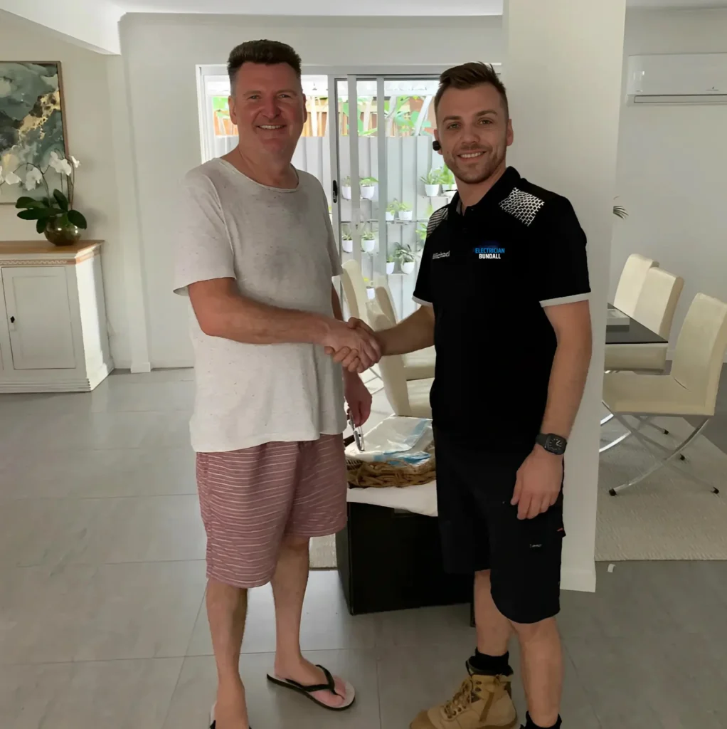 A tradesman in a black uniform shakes hands with a smiling homeowner in casual clothes inside a modern, well-lit home with white walls, tiled floors, and a dining area in the background.